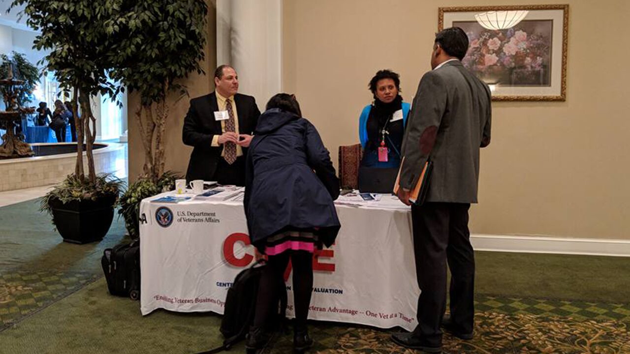 People interacting at an informational booth with a U.S. Department of Veterans Affairs banner, set in a lobby area with plants and artwork.