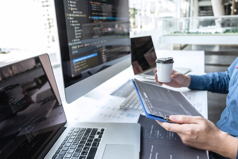 Person holding a clipboard and cup, working on multiple computers with code on screens.